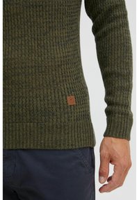 Ciemnozielony sweter wykonany z dzianiny, zdobiony strukturalnym wzorem, z prążkowanymi mankietami oraz małą brązową metką na dolnym brzegu. Tekstura z mieszanki bawełny lub wełny.