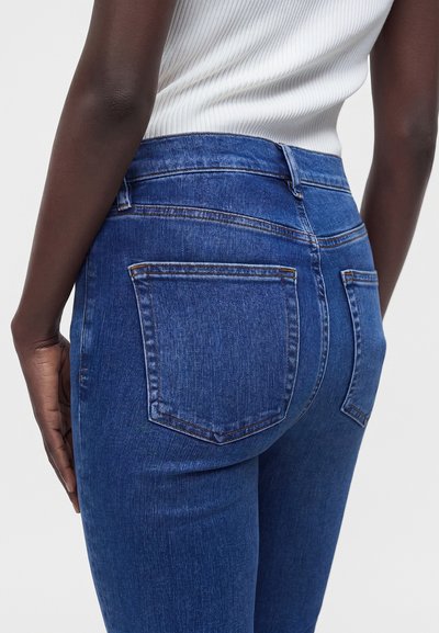 Κοντινό πλάνο ενός ατόμου που φορά μπλε τζιν ψηλόμεσο και λευκό ριμπ πλεκτό αμάνικο τοπ, που δείχνει τις πίσω τσέπες και το πλάι του χεριού.