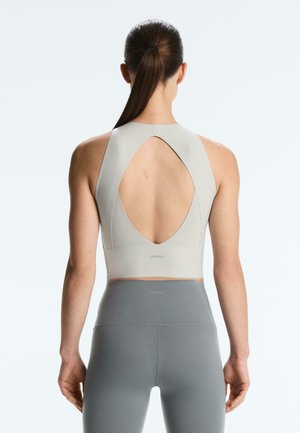 Fekete lófarokkal rendelkező nő, aki világosszürke, ujjatlan, nyitott hátú crop topot és szürke, magas derekú leggingset visel, egyszerű háttér előtt áll.