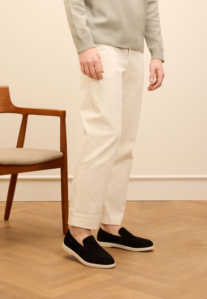 Black suede slip-on shoes with a white sole, paired with cream-colored trousers and a light green sweater, standing beside a wooden chair.