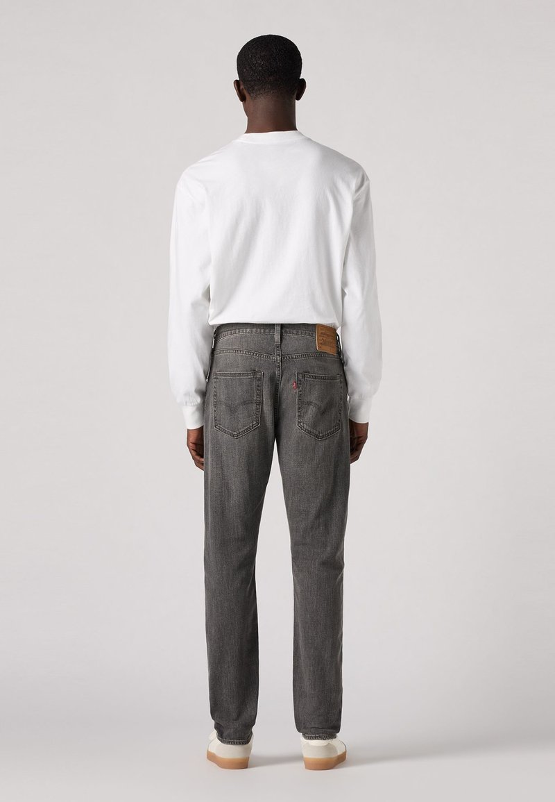 Jean en denim gris à coupe droite, avec une étiquette en cuir marron à la taille et deux poches arrière. Porté avec une chemise blanche à manches longues.