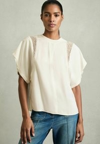 Cremfarbene Bluse mit kurzen Ärmeln, Spitzenverzierungen an den Schultern und lockerer Passform. Kombiniert mit blauen Denim-Jeans im klassischen Schnitt.