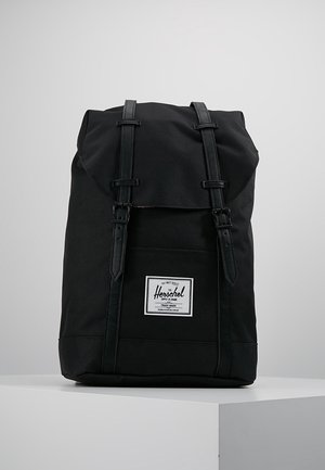 Zwarte Herschel-rugzak met twee riemen en gespen aan de voorkant, een centraal geplaatste logopatch, staand op een wit voetstuk tegen een grijze achtergrond.