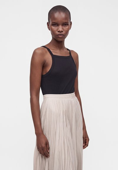 Jeune femme aux cheveux courts portant un top noir sans manches et une jupe plissée beige clair taille haute, debout devant un fond uni.