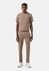 Lysebrun kortærmet t-shirt med lille logo på brystet, kombineret med lysebrune slim fit bukser og hvide atletiske sko.