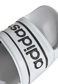 Vit halksandaler med svart Adidas-logotyp. Tillverkade av textilsyntetmaterial med ett tjockt band och vadderad sula.