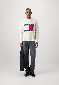 Jersey crema con un diseño de logo en azul marino, rojo y blanco; combinado con jeans grises y zapatos negros; sosteniendo un bolso negro.