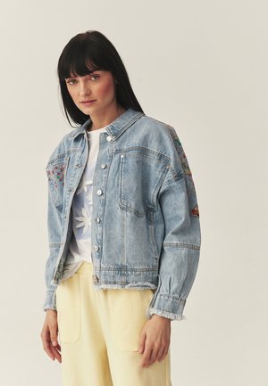 Giovane donna con giacca di jeans azzurra ricamata sopra una camicia bianca floreale e pantaloni giallo pallido, in piedi su uno sfondo semplice.