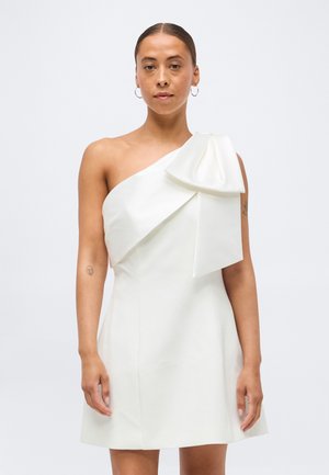 Femme portant une robe blanche asymétrique avec un grand nœud sur l'épaule, debout devant un fond clair uni.