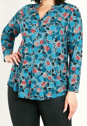 Kvinde iført en turkis blomsterprintet bluse med lange ærmer med blå og røde blomster, kombineret med sorte bukser, hånd på hoften, vist fra nakke til hofter.