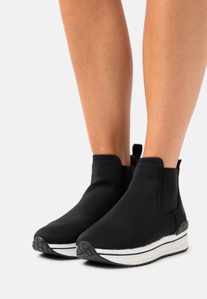 Nohy v černých kotníkových botách na platformě s bílými podrážkami a poutky na bílém pozadí.