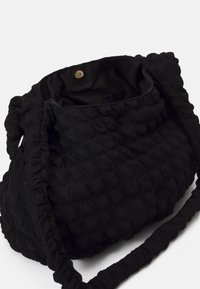 Sac à épaule en tissu noir texturé avec une bandoulière froncée douce et une fermeture à bouton-pression, présenté ouvert avec la doublure intérieure visible.