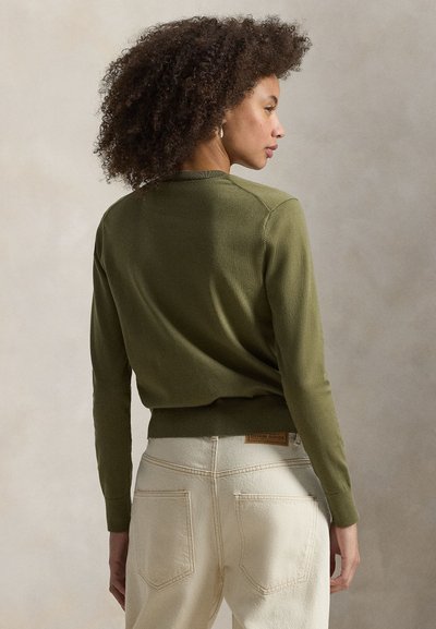 Πλεκτό πουλόβερ ελιάς με στρογγυλή λαιμόκοψη και ριμπ τελείωμα, σε συνδυασμό με τζιν παντελόνι ανοιχτού χρώματος που διαθέτει δερμάτινη ετικέτα στο πίσω μέρος.