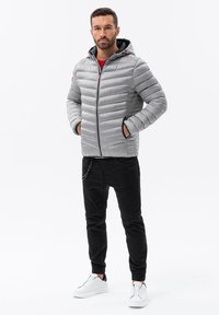 Grå pufferjacka med horisontell sömnad, svarta detaljer och huva. I kombination med svarta joggers och vita sneakers, stående mot en vit bakgrund.
