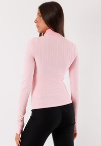 Top a maniche lunghe di colore rosa chiaro a coste con colletto alto. Presenta una silhouette aderente, un motivo testurizzato e maniche affusolate. Pantaloni neri visibili.