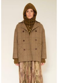 Manteau en laine à double boutonnage en carreaux marron avec de grandes poches plaquées, superposé à un pull olive-texturé, porté avec une capuche assortie.