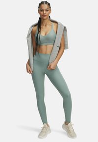 Light green sports bra and high-waisted leggings, paired with a gray hoodie draped over the shoulders; model wears beige athletic shoes.