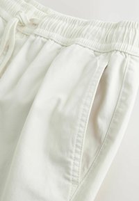 Nærbillede af hvide bukser med elastisk taljebånd med snøre og sidelomme med synlige syningsdetaljer.