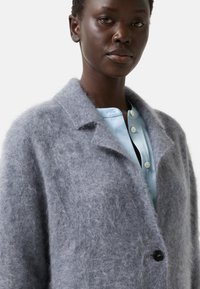 Dark-skinned person wearing a soft, fuzzy gray coat over a light blue buttoned shirt against a plain light background.