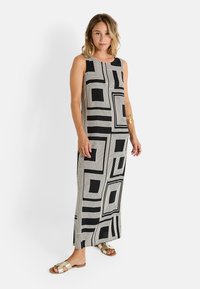 Robe maxi sans manches en noir et blanc avec un imprimé géométrique en carré, fabriquée en tissu lisse. Associée à des sandales métalliques.
