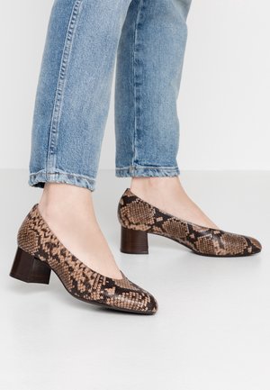 Legs in cuffed blue jeans wearing brown snake print low block heel shoes on a light floor against a plain white wall.