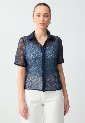 Kobieta ubrana w granatową, koronkową bluzkę na guziki z krótkim rękawem nałożoną na dopasowany top typu camisole, w połączeniu z białymi spodniami, stojąca na jednolitym tle.