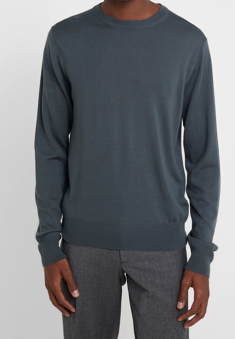 Maglione lungo a maniche nere in morbido tessuto, con collo a giro e polsini e orlo a coste. Indossato con pantaloni grigi a motivo.