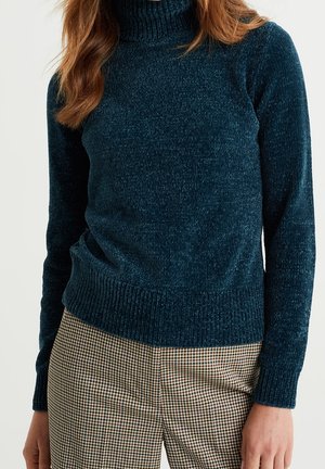 Mörk teal stickad polotröja med ribbade muddar och nederkant. Har ett strukturerat mönster och en figursydd silhuett. Kombinerad med rutiga byxor.