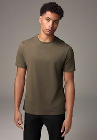 Olivově zelené tričko s krátkým rukávem a kulatým výstřihem, vyrobené z hladkého materiálu, vystavené na modelce na neutrálním pozadí.