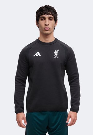 Homme portant un sweat-shirt noir du Liverpool FC et un pantalon de sport bleu sarcelle, debout devant un fond clair uni, faisant face à l’avant.