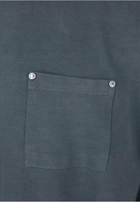 Mørkegrå bomulds t-shirt med en brystlomme med en sølv snapknap og en glat tekstur. Sømdetaljer er synlige omkring lommen.
