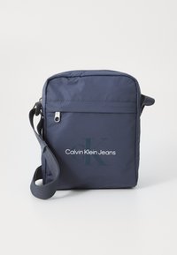 Sötétkék Calvin Klein Jeans keresztpántos táska �állítható pánttal, cipzáras első zsebbel és nyomtatott logóval az elülső panelen.