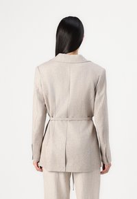 Vaalean beige bleiseri, jossa on jämäkkä muotoilu, yksirivinen, vyöllinen vyötärö ja hienovarainen rakenne. Varustettu lovitettulla kaula-aukolla ja takapuvan tuuletusaukoilla.