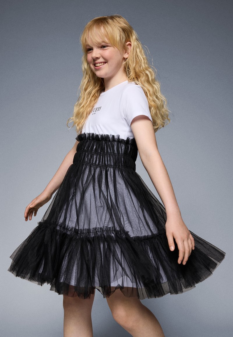 Rochie din tul negru cu un corset ajustat și un fustă stratificată. Cămașă albă cu mânecă scurtă dedesubt cu text. Textură netedă, aspect semi-transparent.