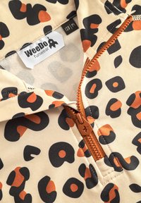 Beige barnjacka med svart och orange leopardmönster, som har en framsida med dragkedja och kopparfärgad hårdvara, samt en mjuk foder.