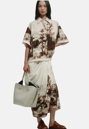 Frau trägt ein beige Kurzarm-Hemdfreihe und passenden Rock mit braunem Baumdruck, hält eine beige Tasche und trägt braune Sandalen.