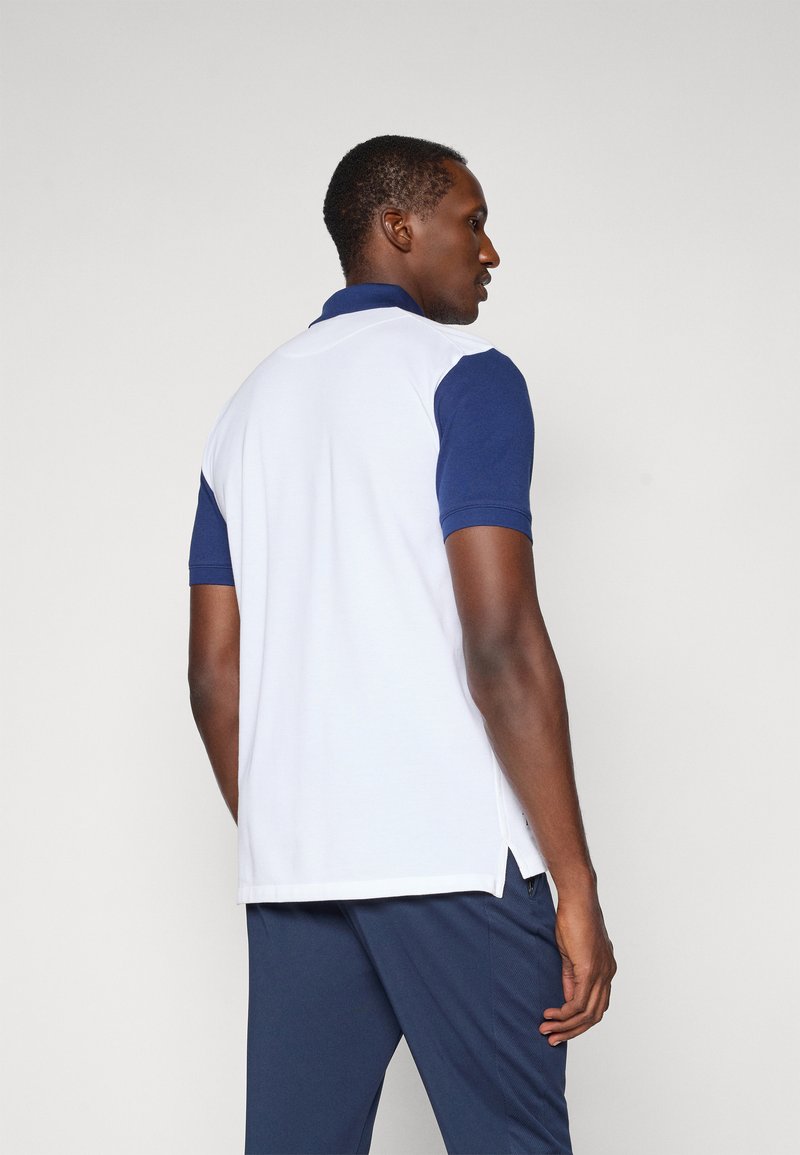 Polo shirt avec un corps blanc et des manches bleu marine, fabriqué en tissu respirant, avec des fentes latérales pour le confort et le mouvement.