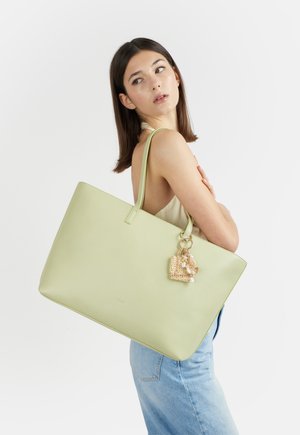 Donna con top senza maniche e jeans che porta una grande borsa tote in pelle verde chiaro con un piccolo charm intrecciato attaccato.