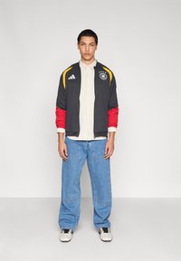 Svart, rød og gul jakke med glidelås, grå skjorte under, blå vide jeans, og svarte og hvite joggesko. Har atletisk design og logo.