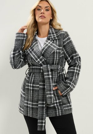Femme portant un manteau à carreaux noir et blanc ceinturé sur un haut blanc et un pantalon noir, posant avec une main dans la poche et l'autre touchant ses cheveux.