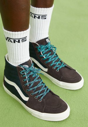 Sneaker alte in suede grigio scuro con accenti turchesi, dotate di lacci turchesi e un logo Vans bianco, posizionate su uno sfondo verde.
