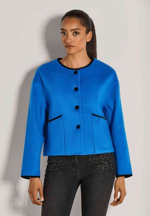 Vrouw met lang haar in een staart, gekleed in een helderblauwe knoopjasje met zwarte afwerking en zwarte jeans met kleine rhinestones.