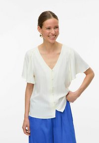 Vit blus med kort ärm och djup V-ringning, vertikala ränder och knäppning. Matchad med blå, löst sittande byxor.