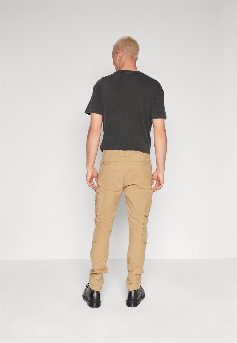 Khaki Cargo-Hosen mit Seitentaschen und schmaler Passform, aus Baumwollstoff. Getragen mit einem lässigen schwarzen T-Shirt und schwarzen Schuhen.