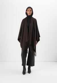 Brauner Poncho aus strukturiertem Strick mit einem Fransensaum. Kombiniert mit einem schwarzen Rollkragenpullover, schwarzen weit geschnittenen Hosen und schwarzen spitzen Stiefeln.