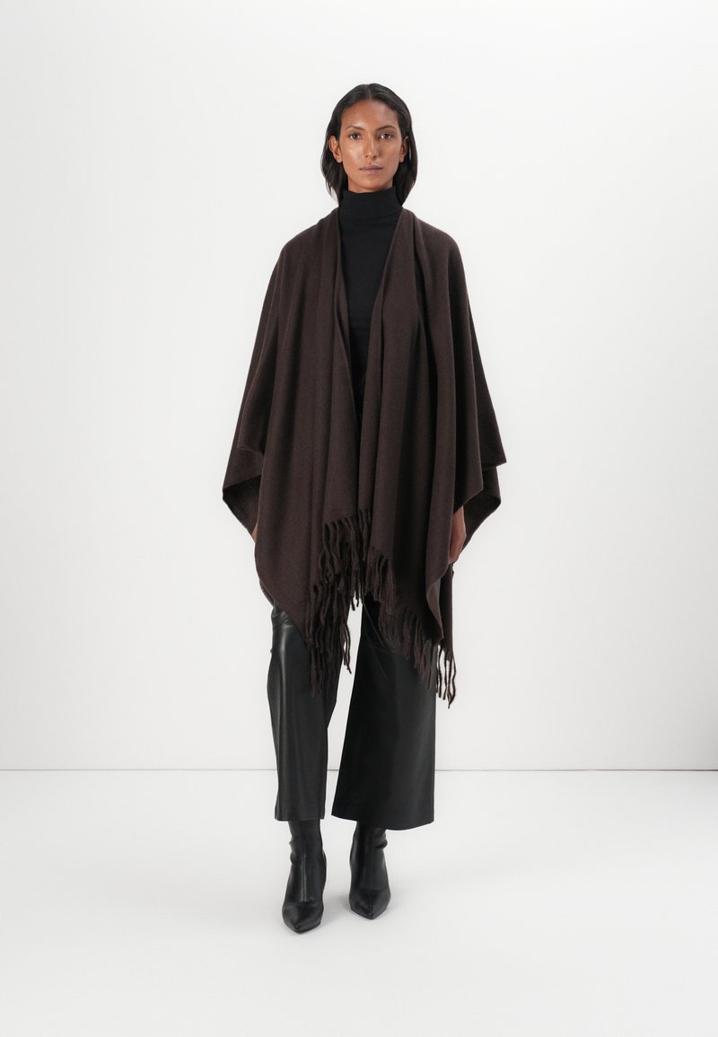 Brauner Poncho aus strukturiertem Strick mit einem Fransensaum. Kombiniert mit einem schwarzen Rollkragenpullover, schwarzen weit geschnittenen Hosen und schwarzen spitzen Stiefeln.