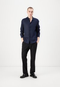 Camicia button-up in lino blu scuro con colletto alto, maniche lunghe e pantaloni plissettati neri. Il modello indossa scarpe nere. Design minimalista.