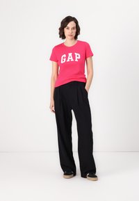 Lys pink t-shirt med hvid "GAP" logo, parret med løse sorte bukser. Modellen står foran en ensfarvet lys baggrund.