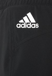Tela negra con el logo de Adidas en blanco impreso cerca de una cintura elástica, mostrando parte de la costura de un bolsillo.
