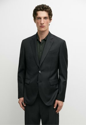 Junger Mann mit lockigem Haar, der eine dunkle Anzugjacke über einem dunklen Hemd mit Knöpfen trägt, vor einem schlichten weißen Hintergrund stehend.